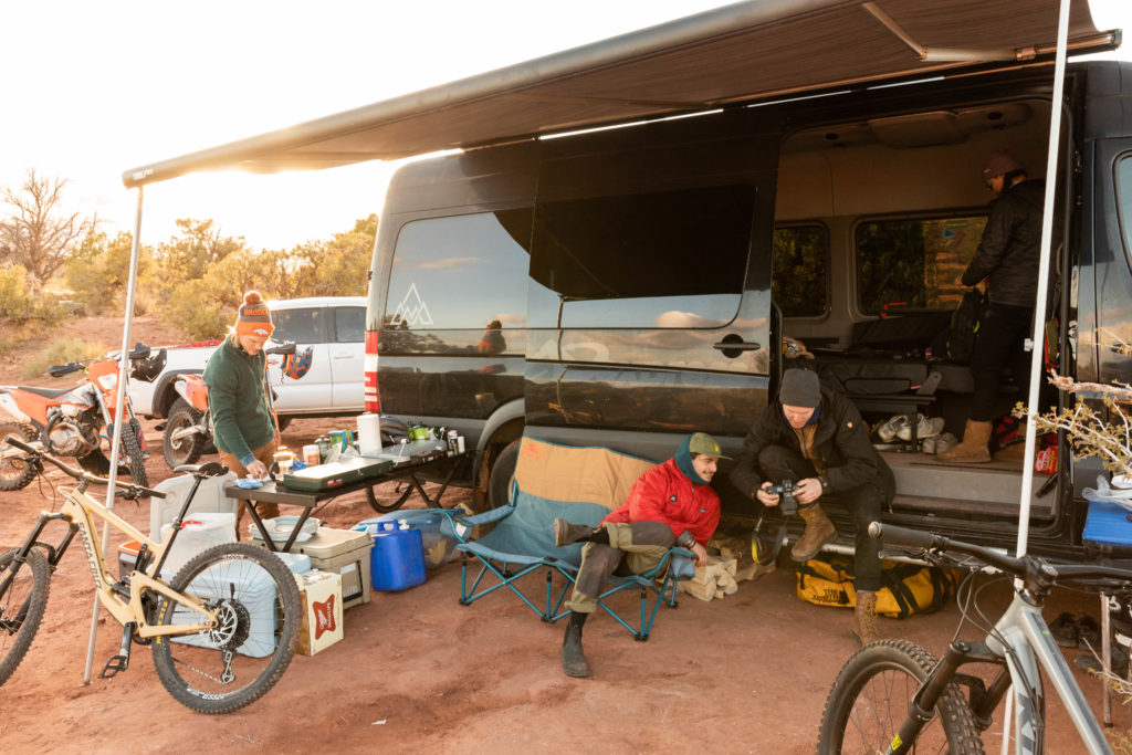 People lounging under an awning around and inside of a MileMarker Campervan with food being made and bikes set aside