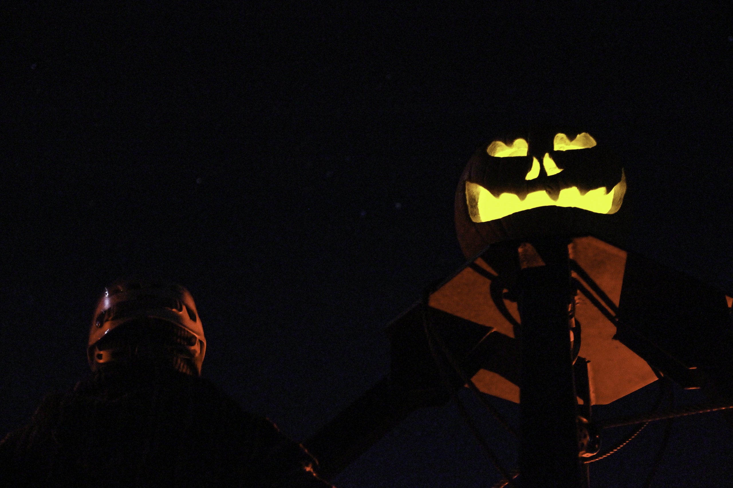 Person with a helmet standing beside a wood post with a jack-o-lantern and reflective vest under the stars